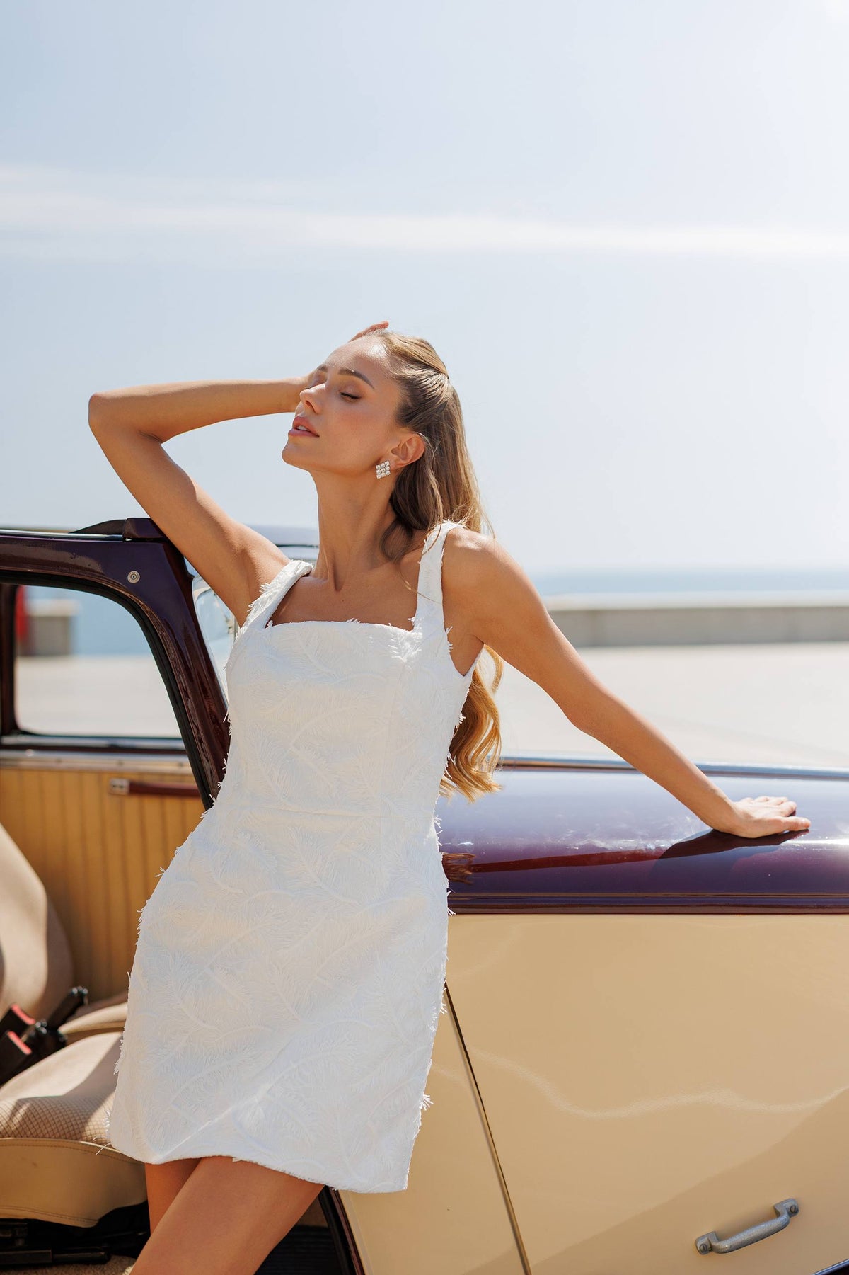 Woman in a white dress standing next to a vintage car with a clear sky background