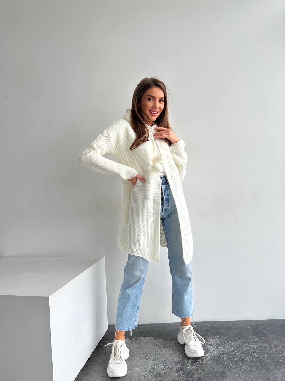 Woman wearing a white cardigan, blue jeans, and white sneakers in a minimalistic setting.