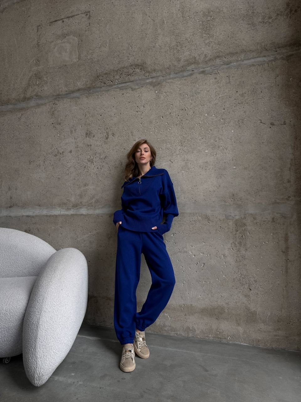 Person wearing a blue outfit standing against a textured wall with a modern chair.