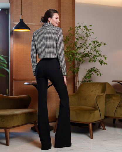 Woman in a checkered shirt and black flared pants standing in a modern room with greenery.