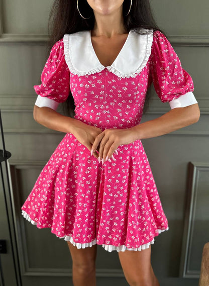 Woman wearing a pink dress with white floral pattern and collar against a neutral background