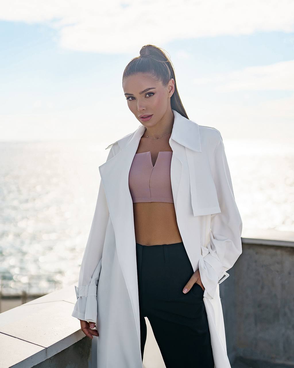Woman wearing a white coat over a pink top and black pants, standing outdoors with water in the background.