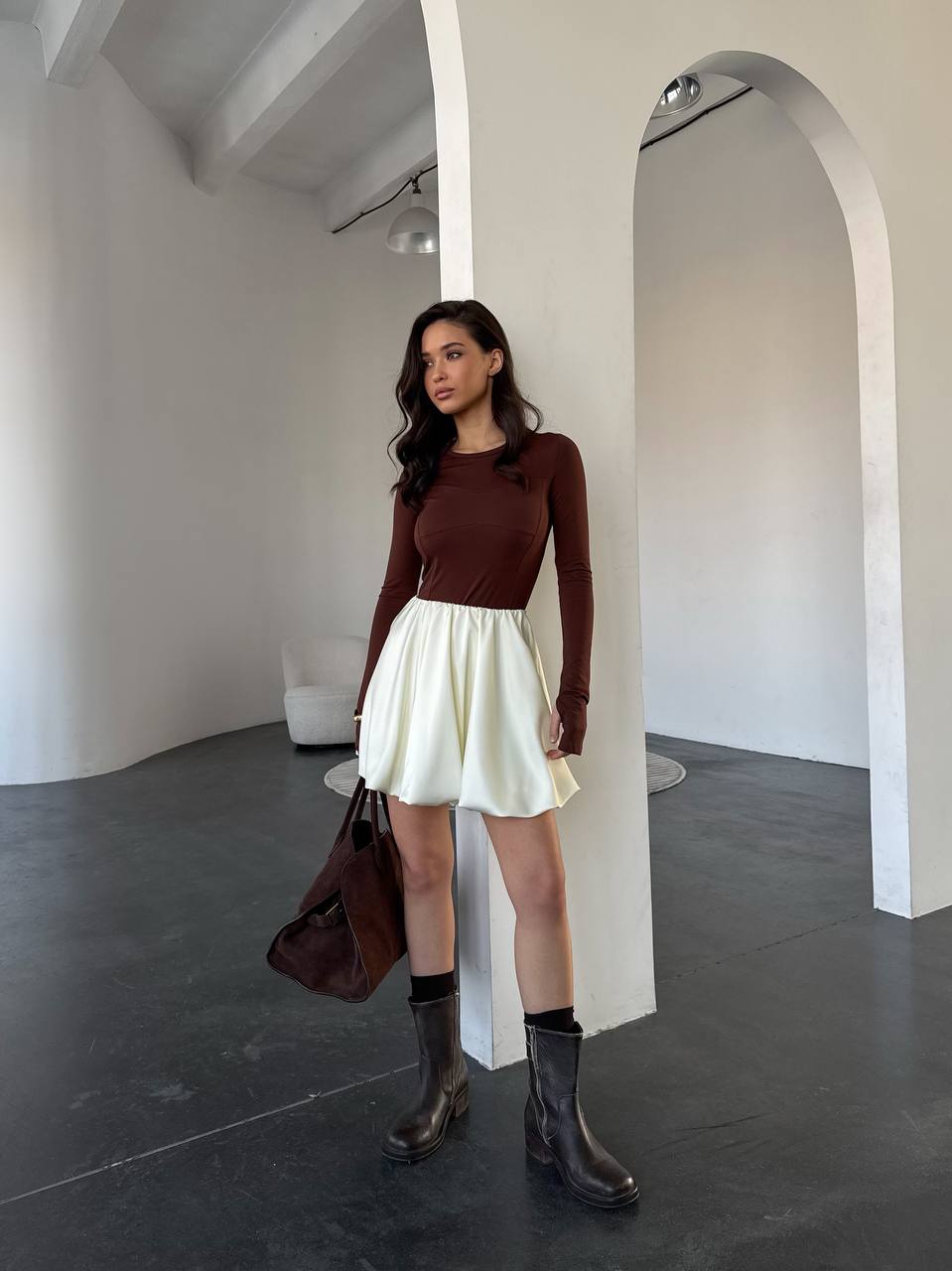Woman in a brown top and white skirt standing in a minimalistic room.