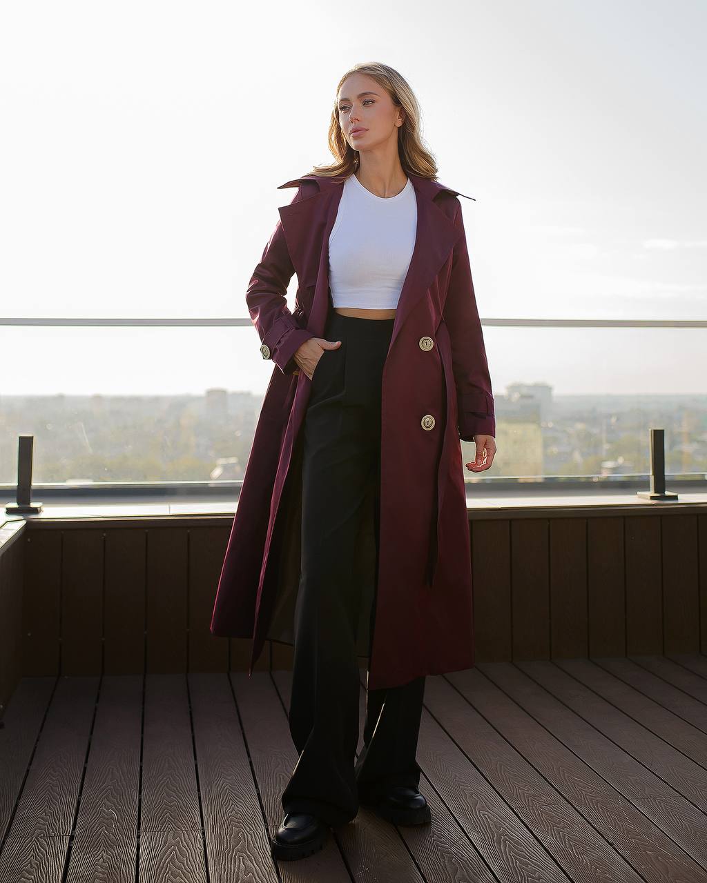 Woman wearing a long burgundy coat with a white top and black pants on a wooden deck.