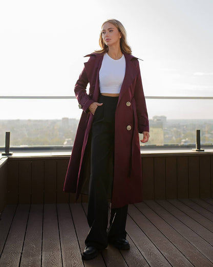 Woman wearing a long burgundy coat with a white top and black pants on a wooden deck.