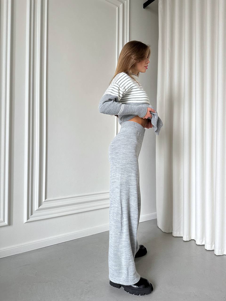 Woman in a gray outfit standing in a room with white walls and curtains.