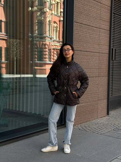 Person wearing a dark quilted jacket and light blue jeans standing in front of a modern building.