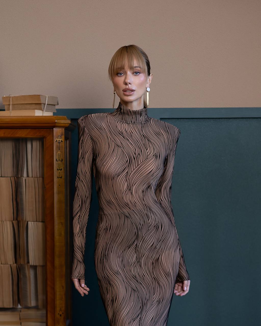 Woman wearing a textured dress standing against a wall with a bookshelf.