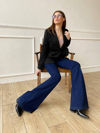 Woman sitting on a chair wearing a black jacket, blue jeans, and black shoes in an elegant room.