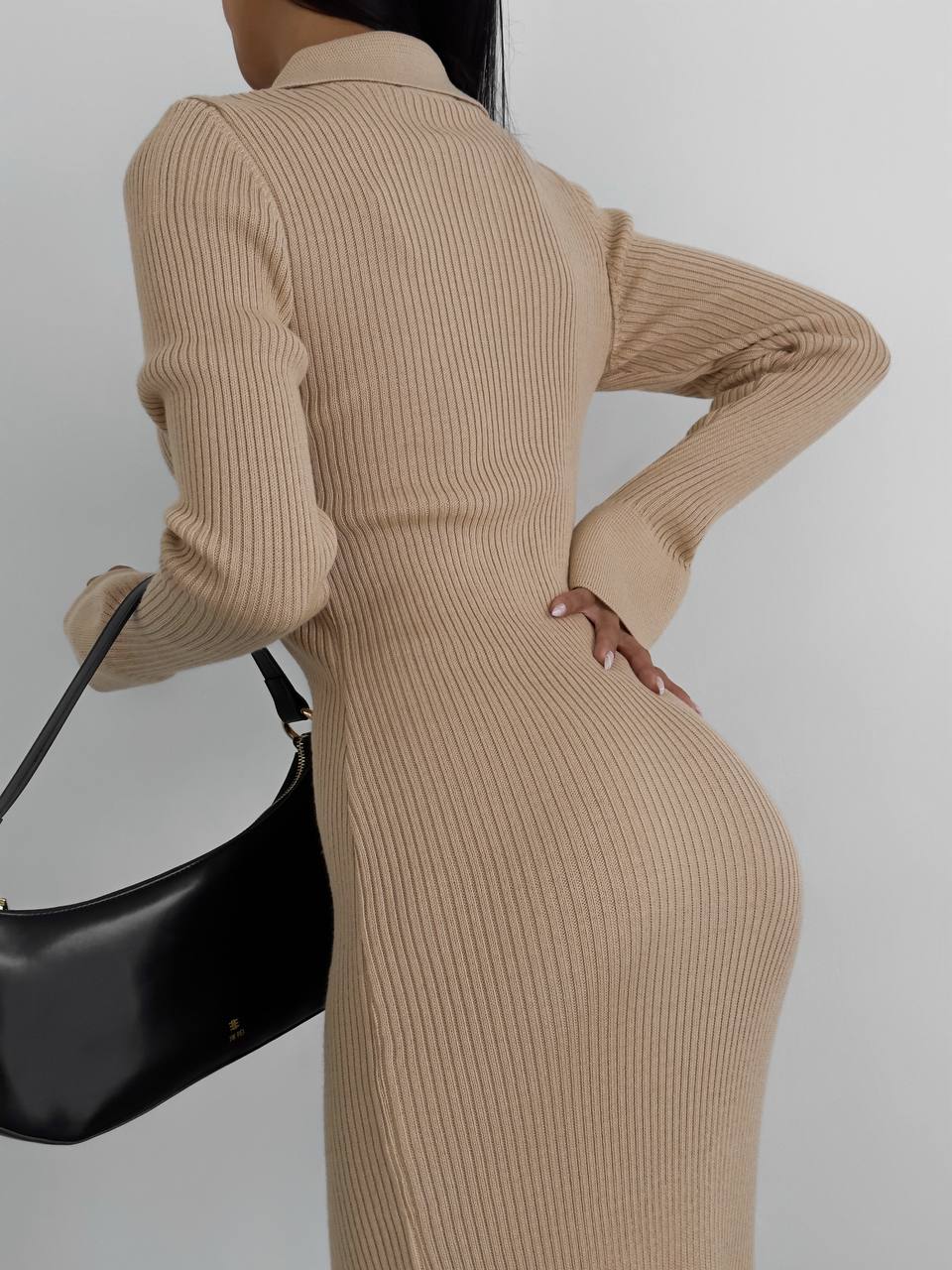 Person wearing a beige ribbed dress holding a black handbag against a plain background