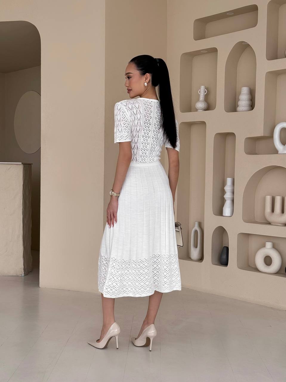 Woman in a white lace dress standing in a modern interior setting with shelves.