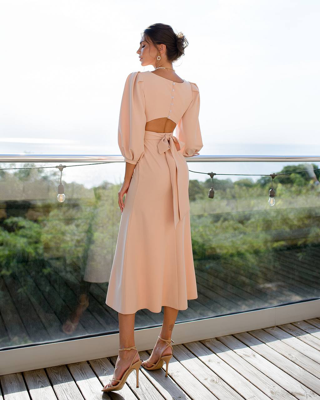 Woman in a beige dress standing on a wooden deck with a glass railing.