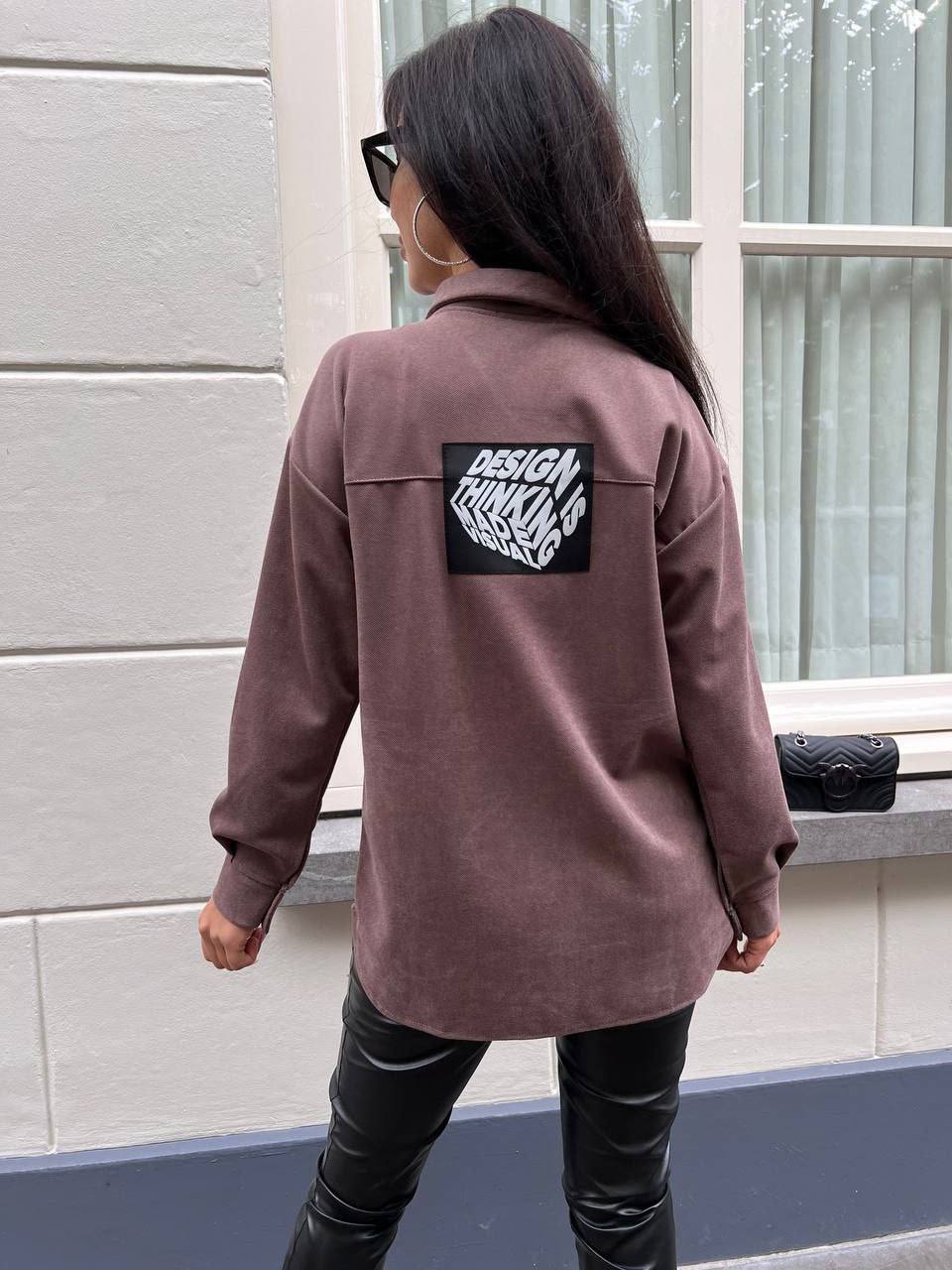 Person wearing a brown jacket with a patch on the back, standing in front of a window.