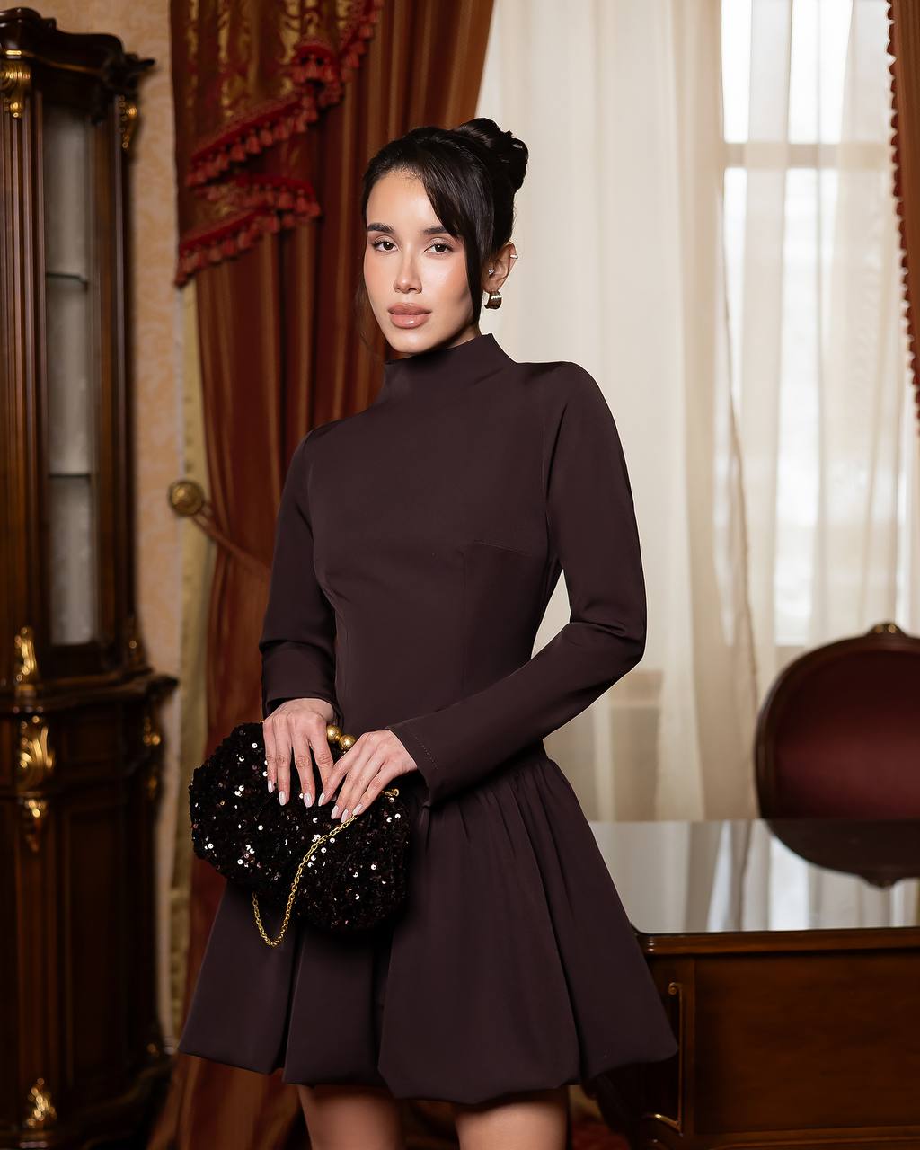 A woman stands indoors wearing a fitted long-sleeve turtleneck dress in very dark plum-brown, holding a black embellished clutch. The room has warm brown drapery, a wooden cabinet, and sheer curtains.