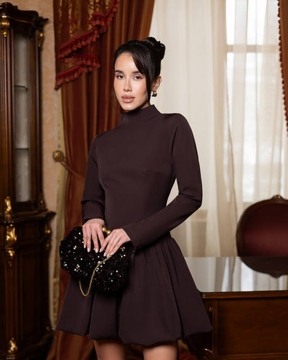 A woman stands indoors wearing a fitted long-sleeve turtleneck dress in very dark plum-brown, holding a black embellished clutch. The room has warm brown drapery, a wooden cabinet, and sheer curtains.