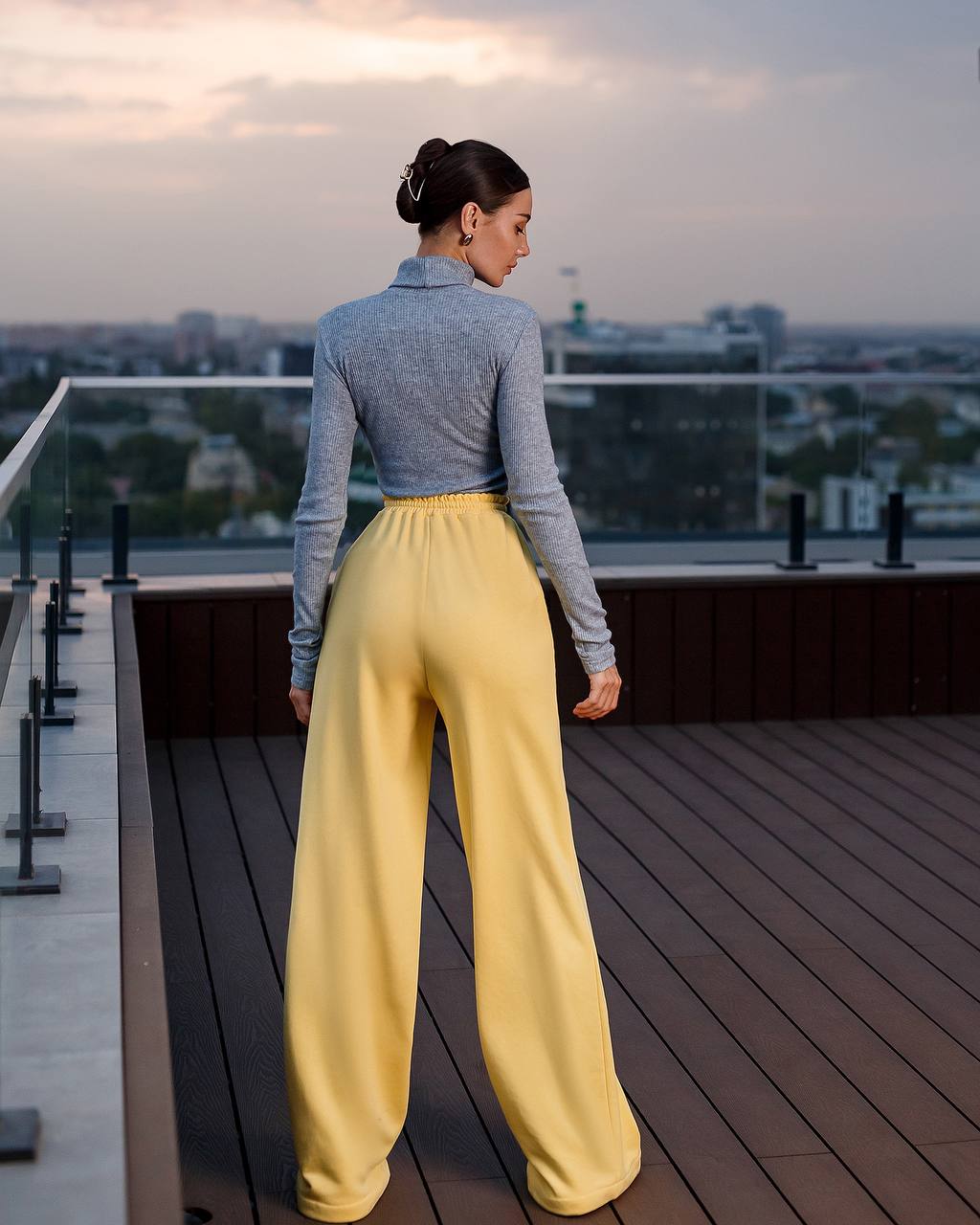 Woman in yellow pants and gray top standing on a rooftop deck with cityscape view.