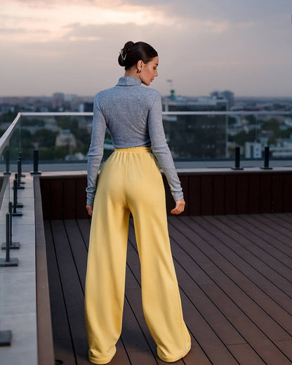 Woman in yellow pants and gray top standing on a rooftop deck with cityscape view.