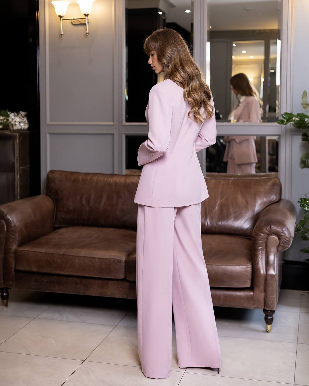 Woman in a pink suit standing in a room with a brown leather couch.