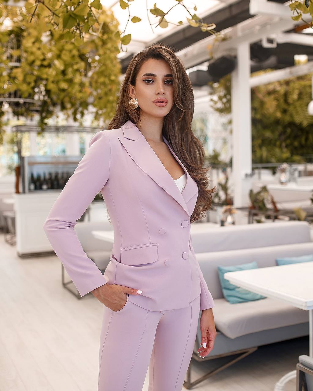 Woman in a lavender suit standing in an outdoor setting with greenery and furniture.