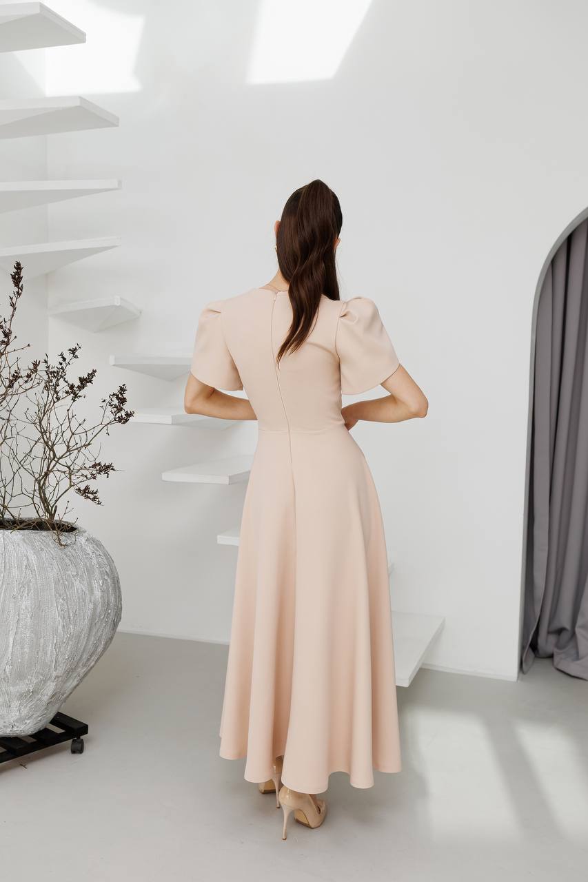 Woman in a beige dress standing in a minimalistic room with white walls and a gray curtain.
