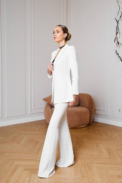 Woman in a white suit standing in a room with wooden flooring and a brown chair.