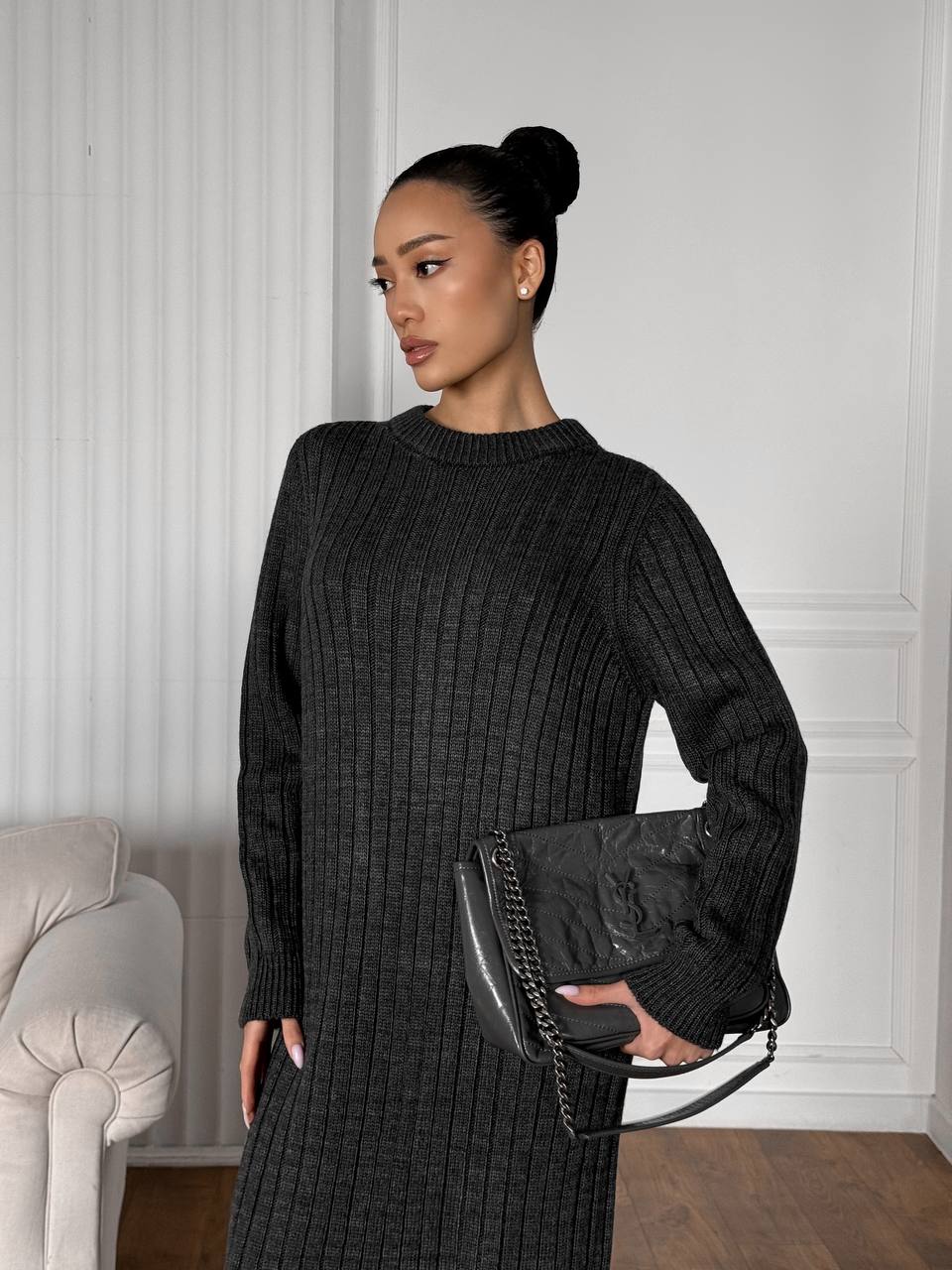 Woman wearing a black ribbed sweater and holding a matching handbag in a room with white walls and a gray chair.