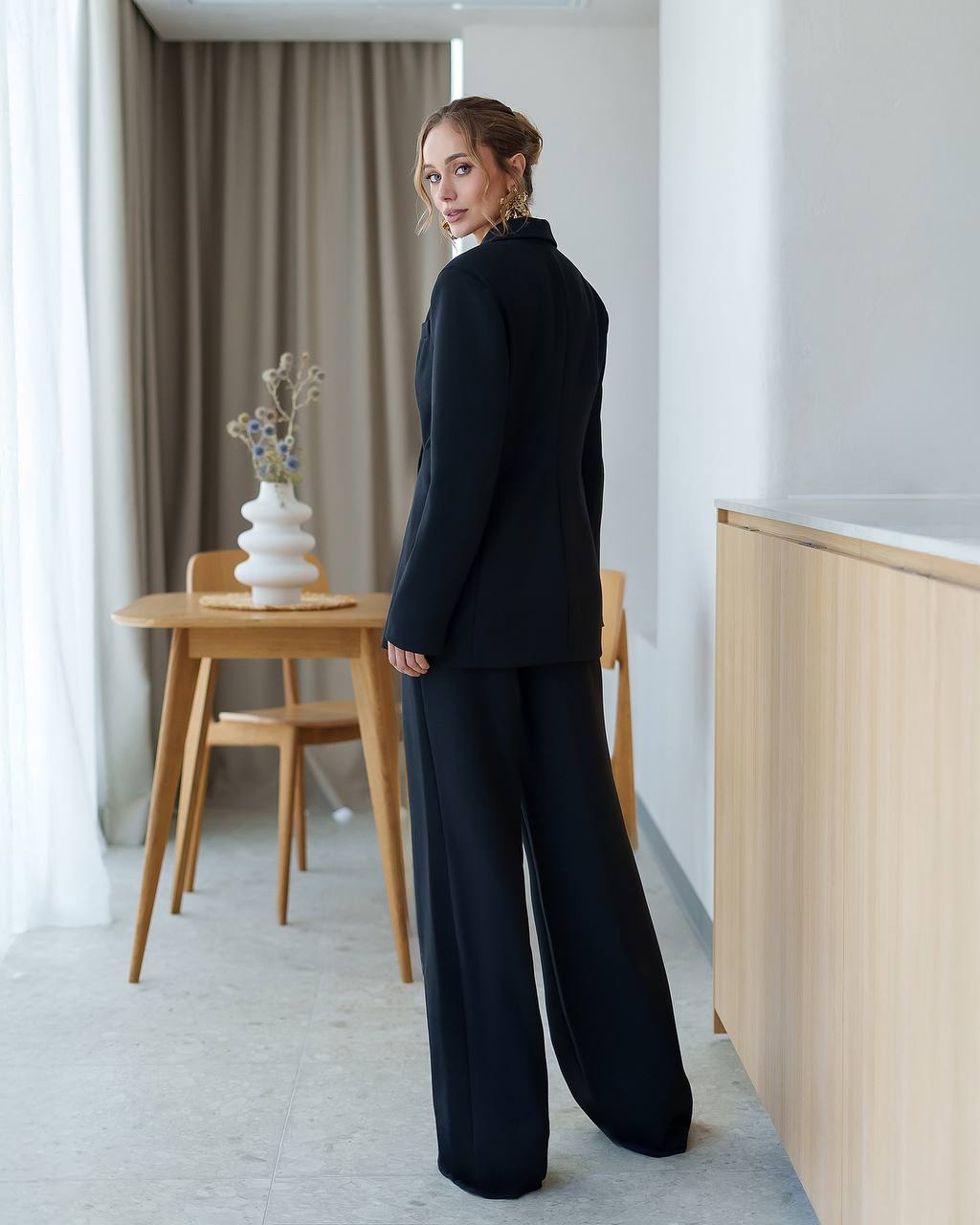 Woman in a black outfit standing in a modern room with wooden furniture and white walls.