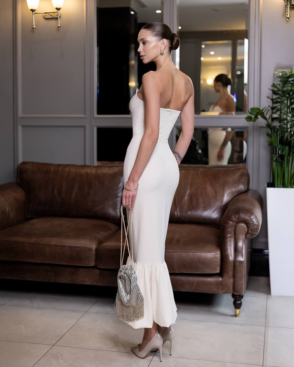 Woman in a white dress standing in a room with a brown leather couch and a mirror.