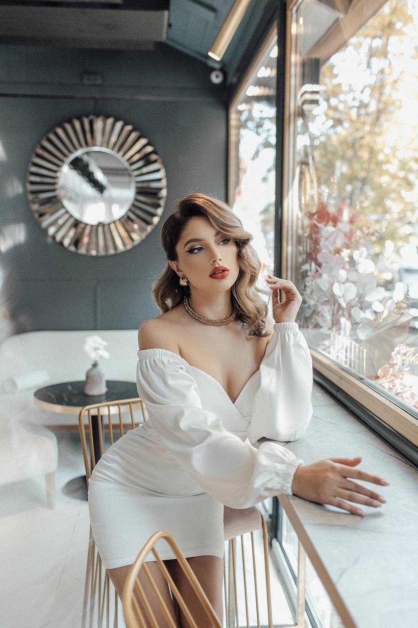Woman in a white dress sitting by a window in a stylish interior.