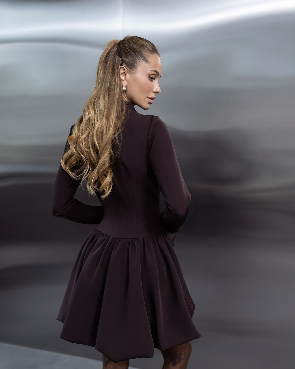 A woman with long blonde hair in a high ponytail is shown from the back wearing a dark plum long-sleeve dress with a flared skirt, set against a dark gray, abstractly textured studio background.