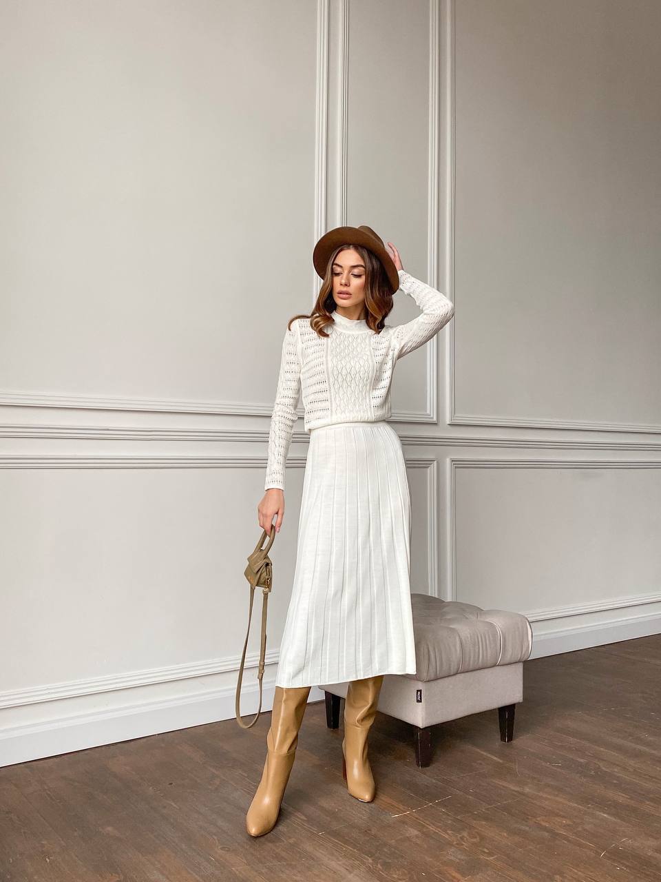 Woman in a white outfit with beige boots standing against a neutral wall.