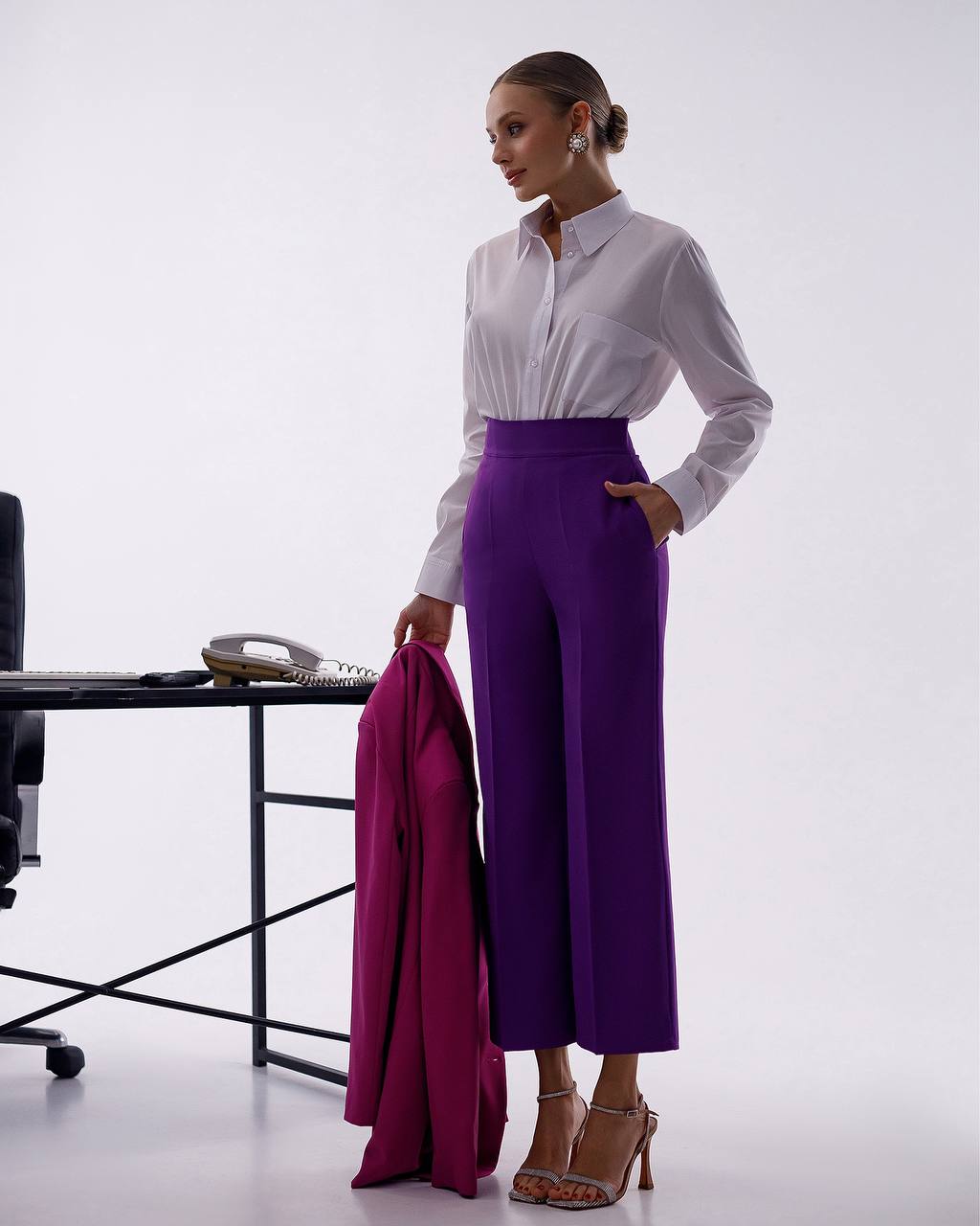 Woman in a white blouse and purple pants standing next to a desk with a phone on it.