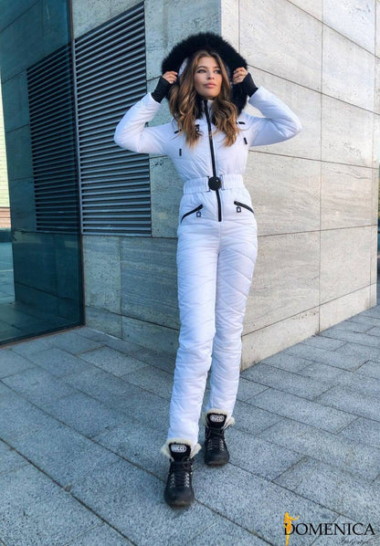 Woman in a white snowsuit with a black fur hood standing against a modern building.