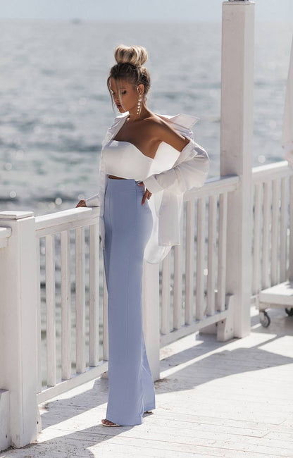 Woman in a white top and light blue pants standing on a wooden deck with ocean view.