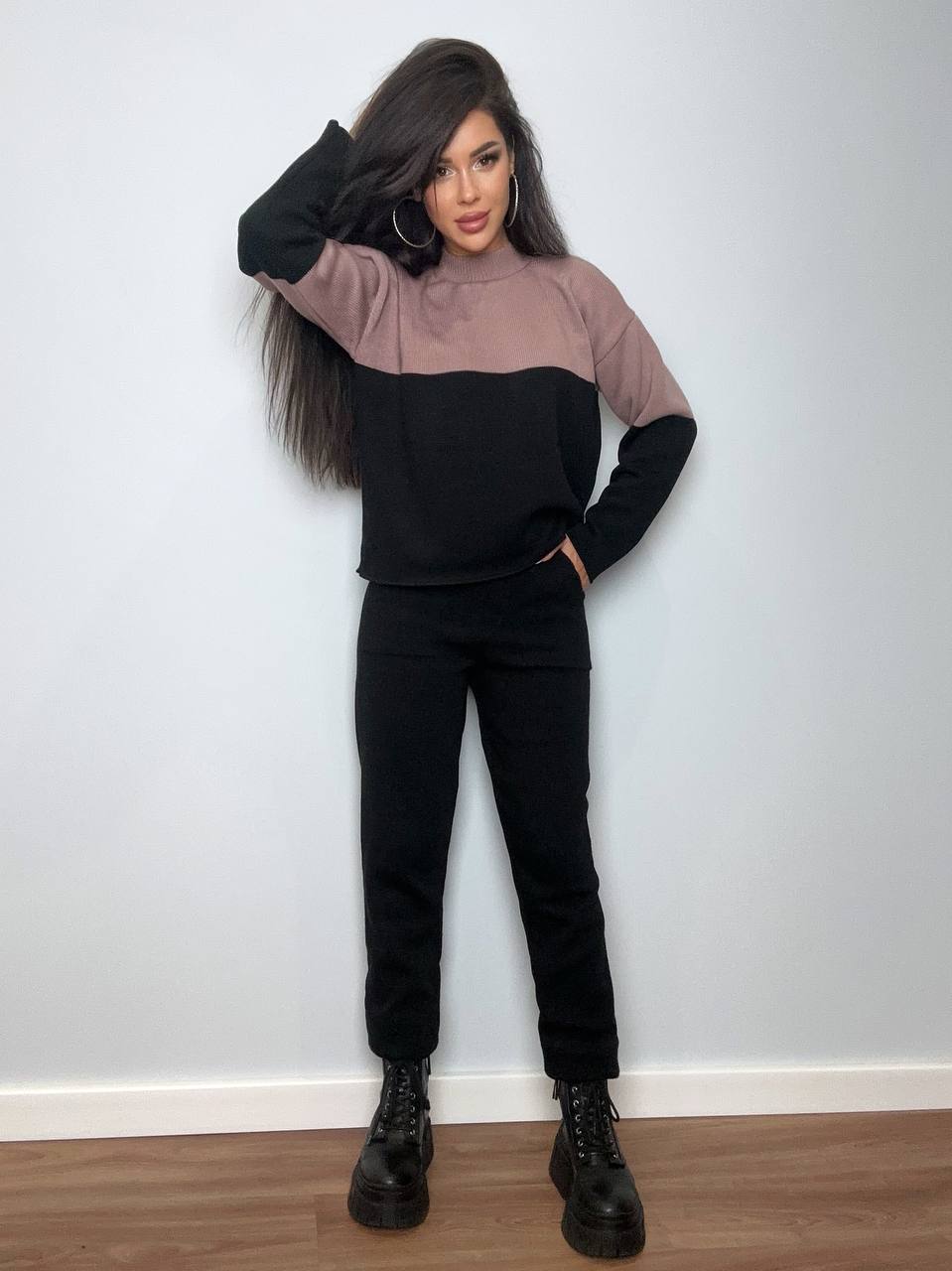 Woman wearing a black and brown color-blocked sweater and black pants against a white wall.