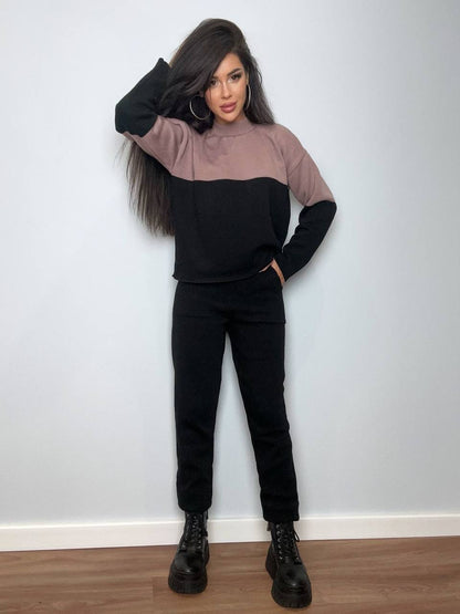 Woman wearing a black and brown color-blocked sweater and black pants against a white wall.
