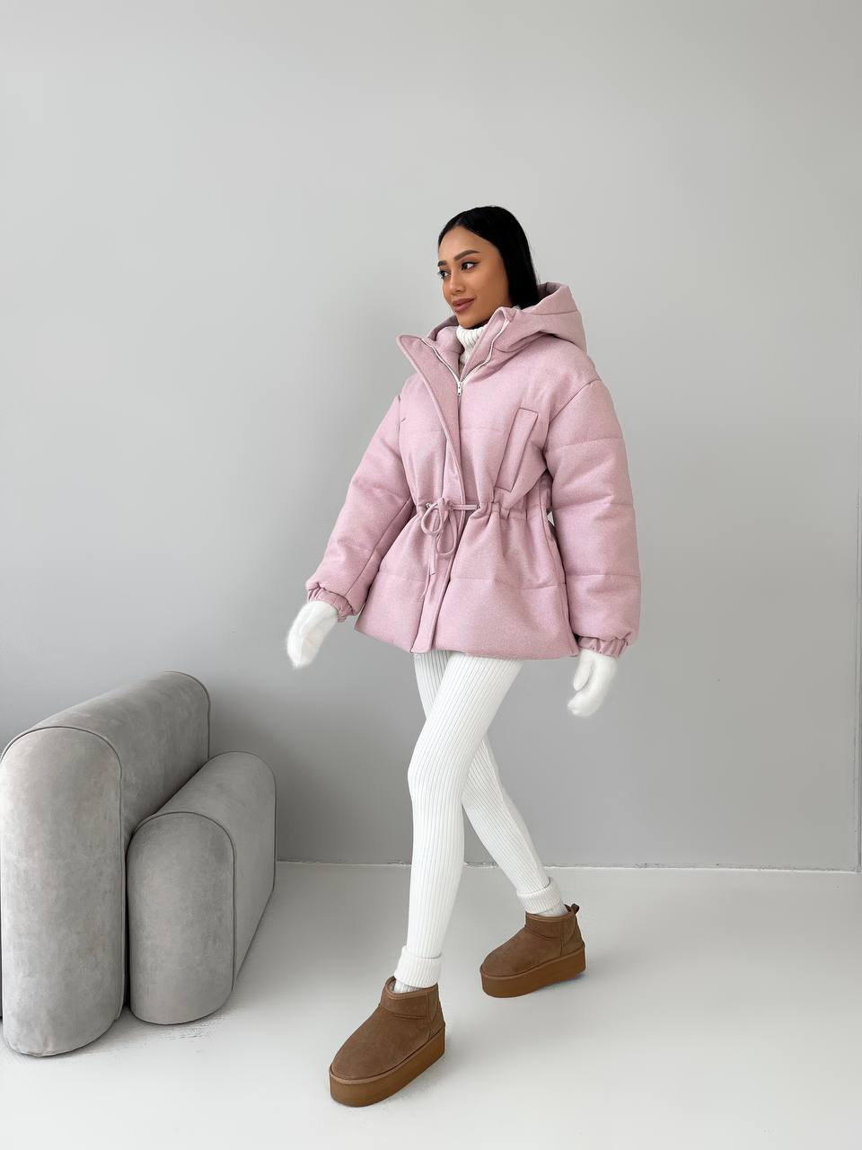 Person wearing a pink puffer jacket with a hood, white leggings, and brown boots in a minimalistic setting.