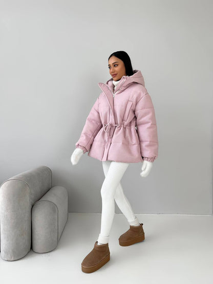 Person wearing a pink puffer jacket with a hood, white leggings, and brown boots in a minimalistic setting.