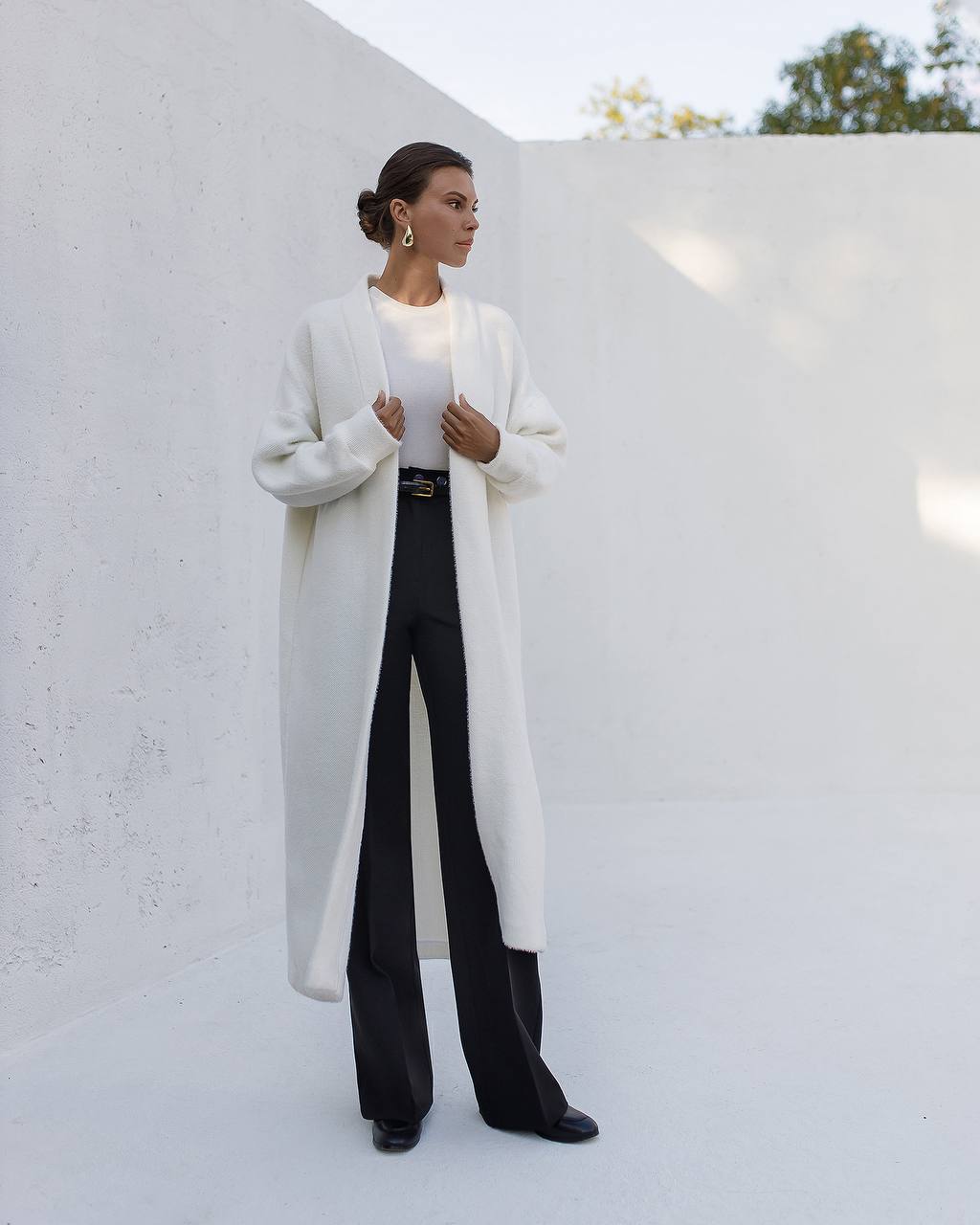Woman wearing a long white coat over a black outfit against a white wall.