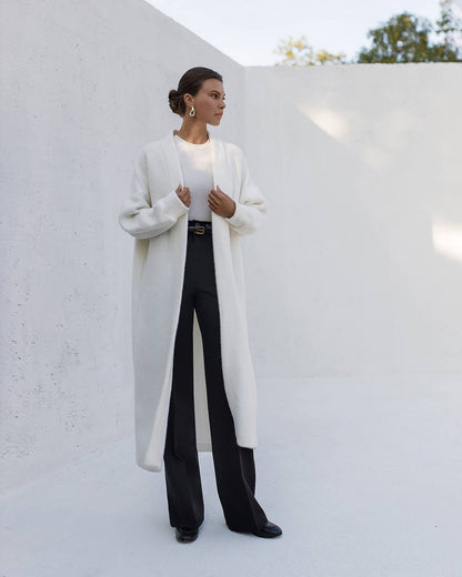 Woman wearing a long white coat over a black outfit against a white wall.