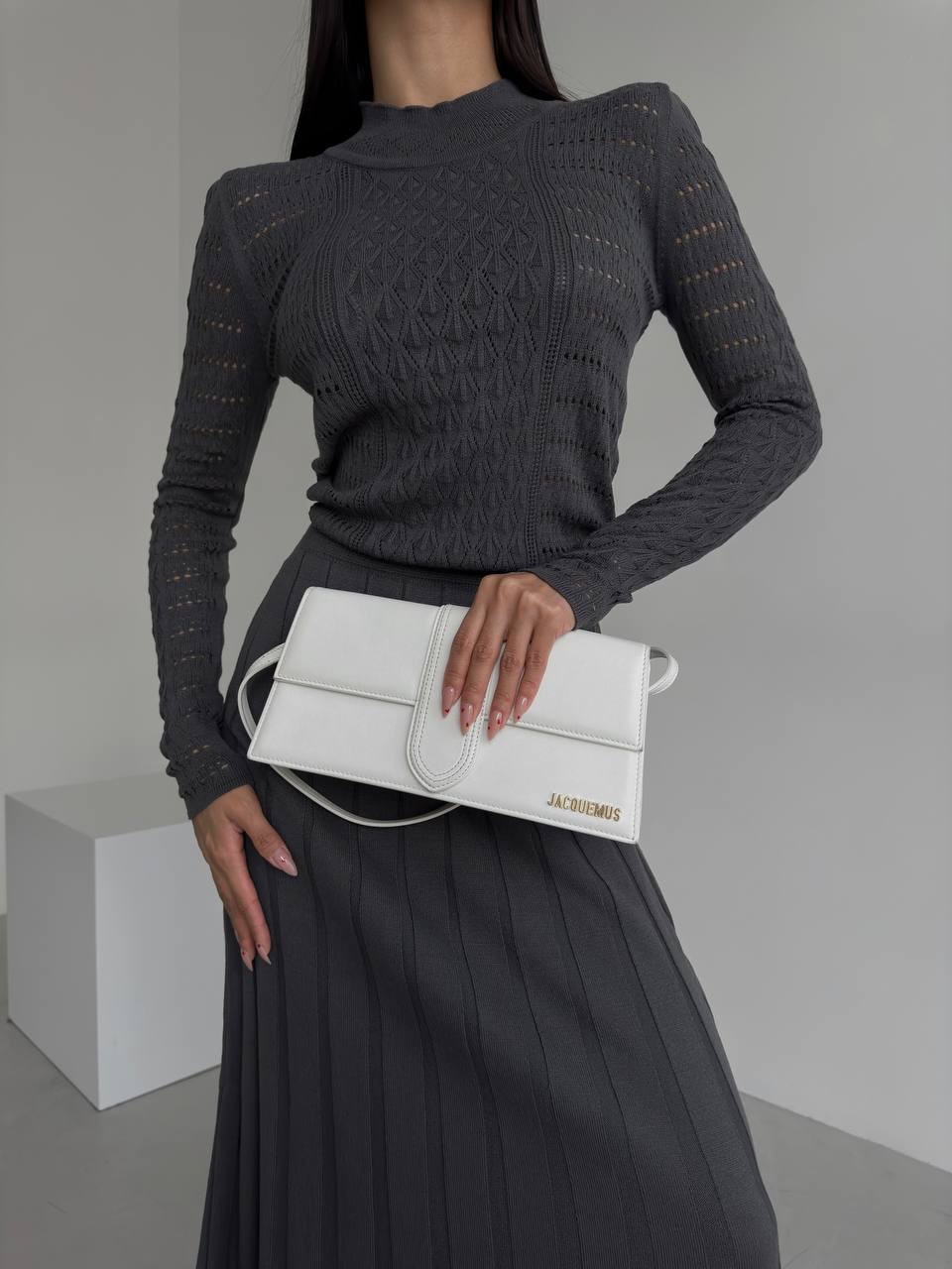 Person wearing a dark gray sweater and holding a white handbag with 'JACQUEMUS' branding.