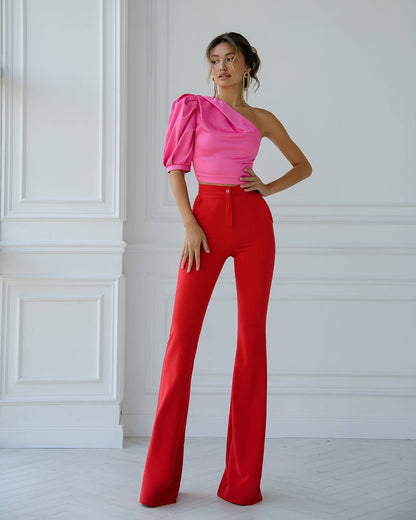 Woman wearing a pink one-shoulder top and red pants against a white wall.