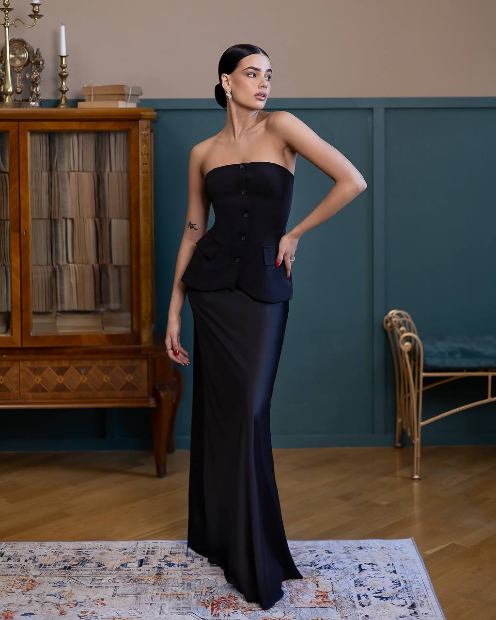 A woman models a strapless black satin gown with a structured peplum-style bodice and a fitted floor-length skirt in an elegant room with dark teal paneling and wood floors.