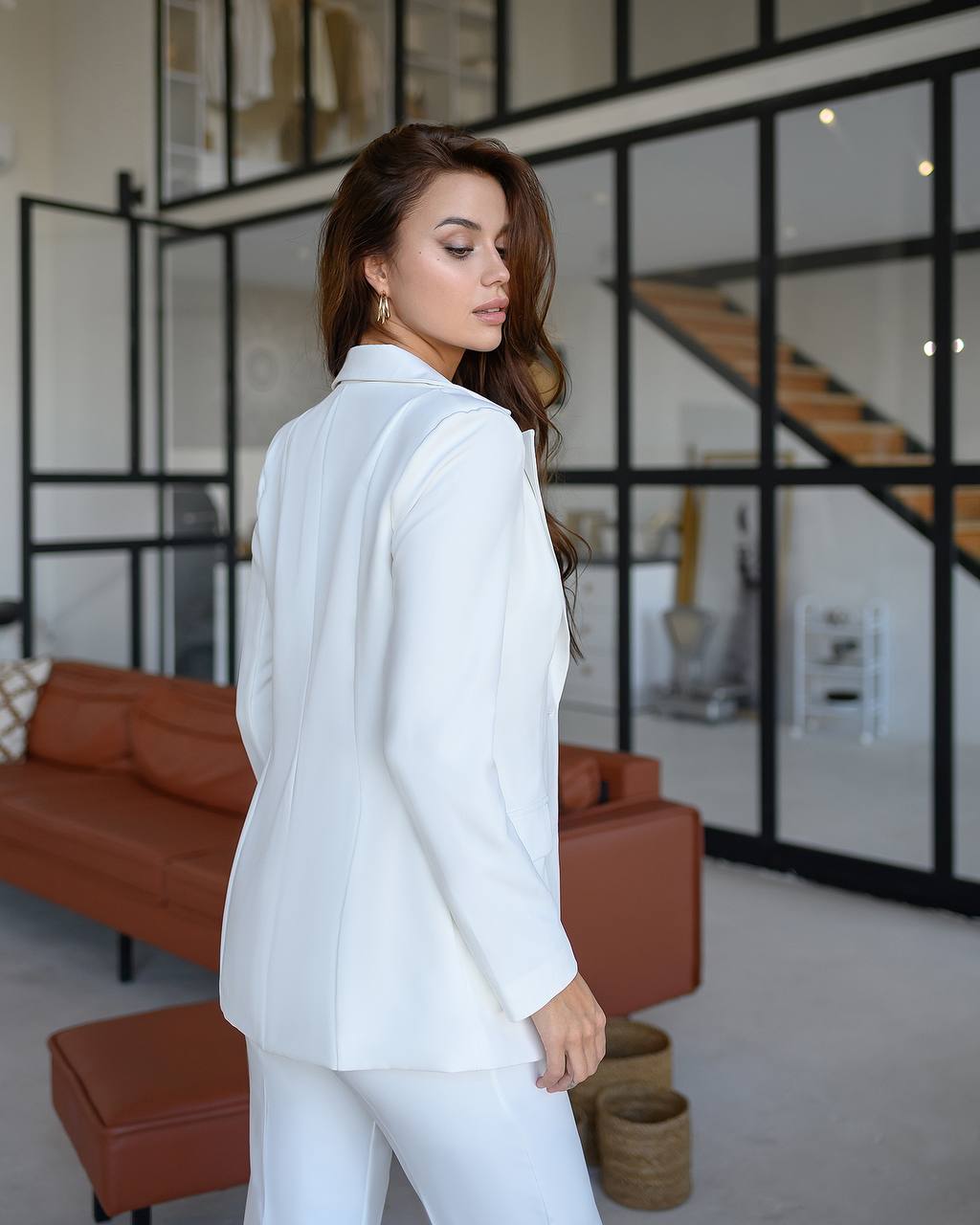 Woman in a white suit standing in a modern living room with a brown sofa and glass walls.