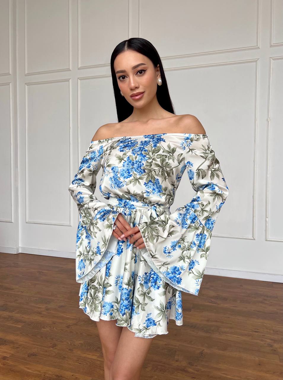 Woman wearing a floral off-shoulder dress against a white paneled wall.