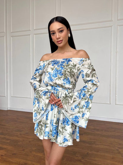 Woman wearing a floral off-shoulder dress against a white paneled wall.