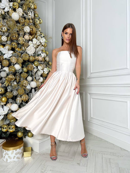 Woman in a white dress standing next to a decorated Christmas tree in a room with white walls.