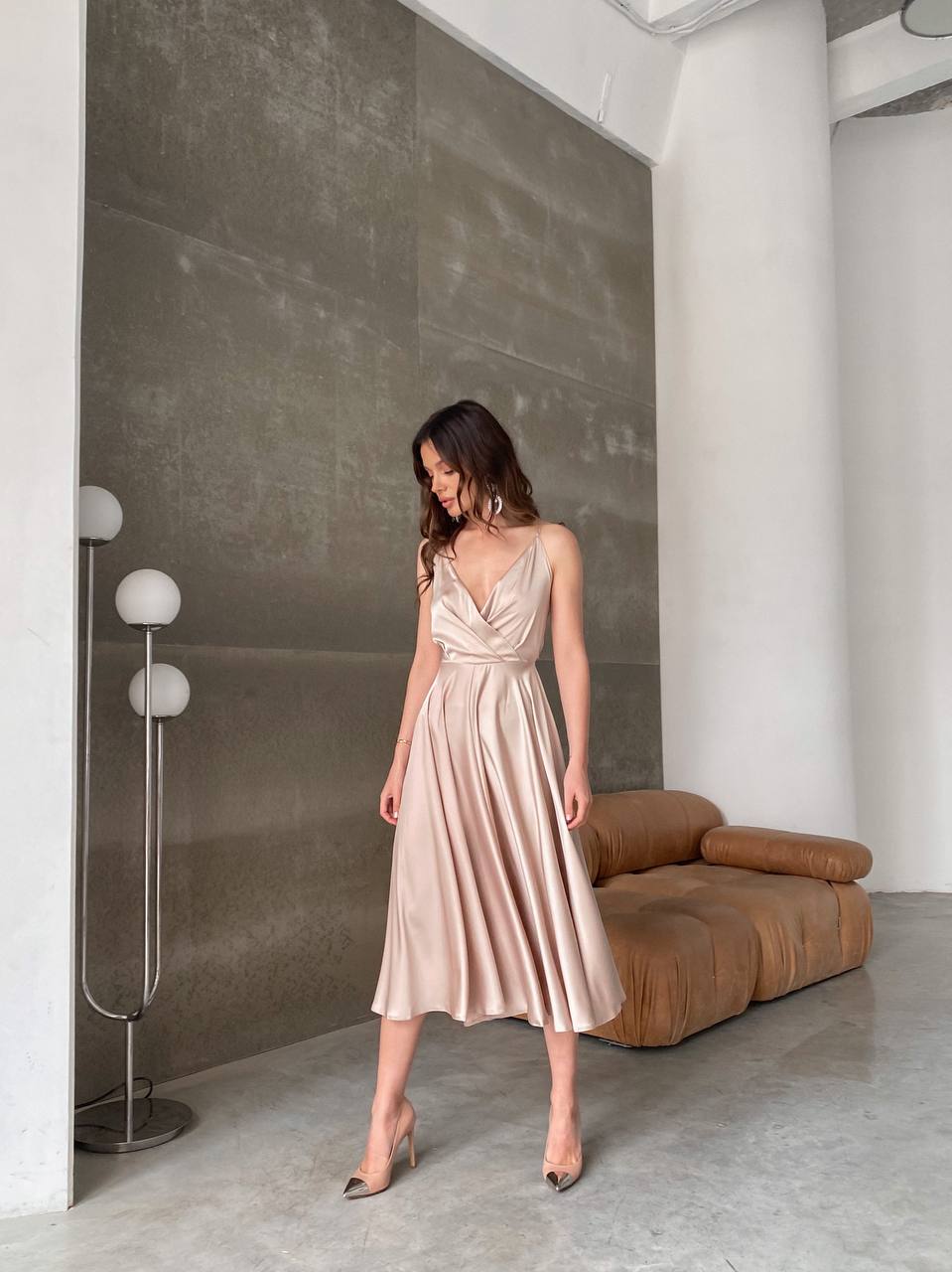 Woman in a beige dress standing in a modern room with a brown sofa and gray walls.