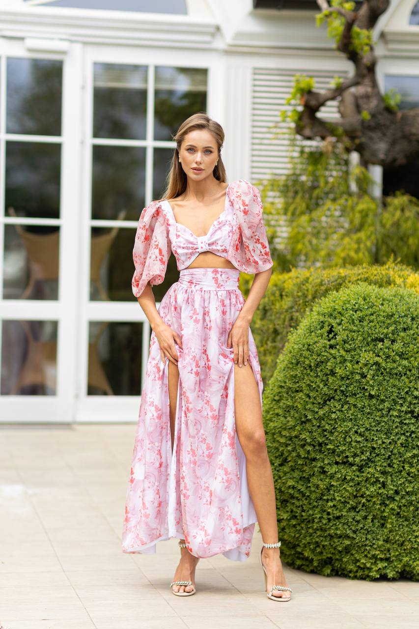 Woman in a pink floral dress standing outdoors with greenery and a building in the background