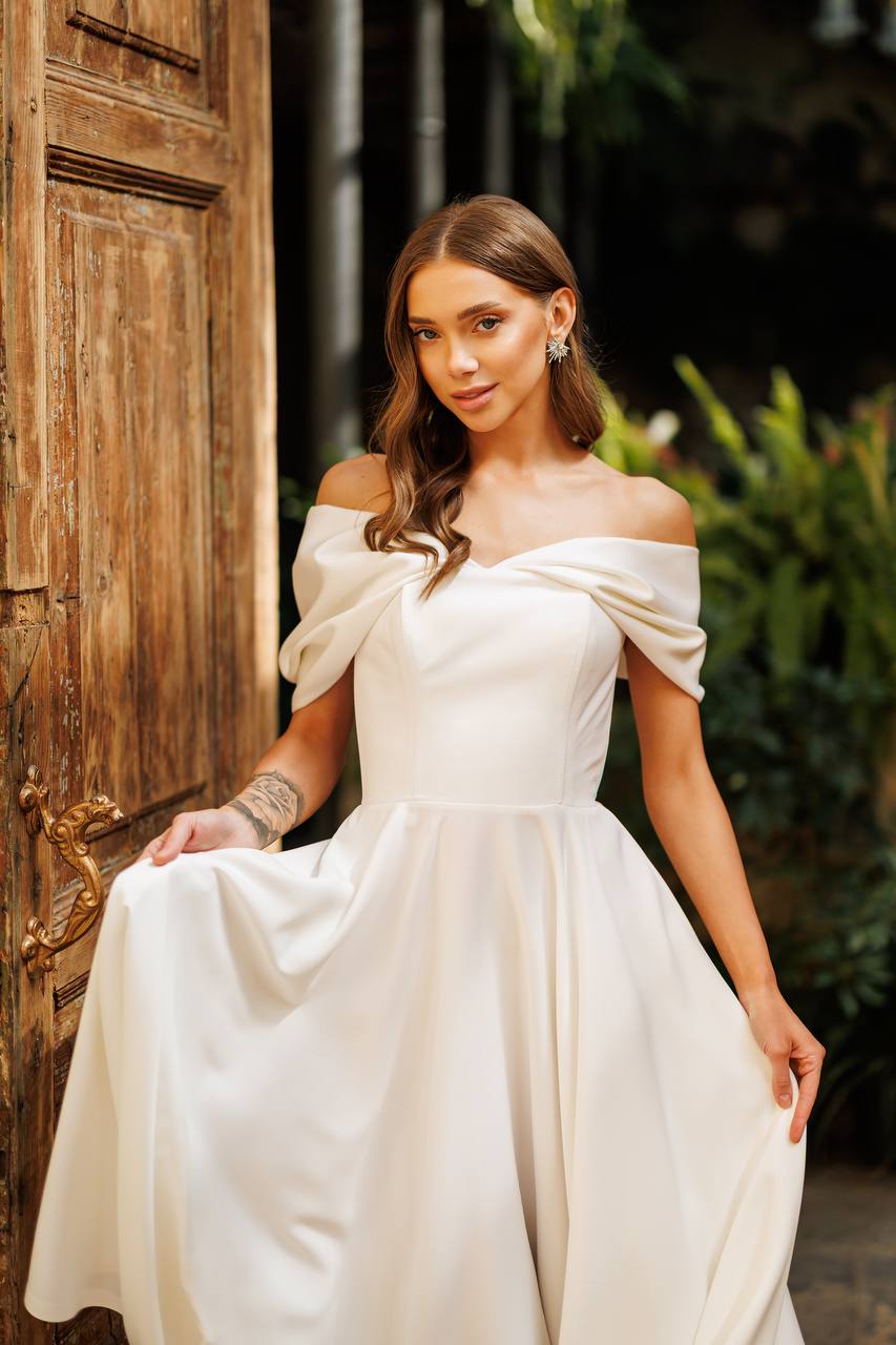 Woman in a white off-shoulder dress sitting outdoors near a wooden door.